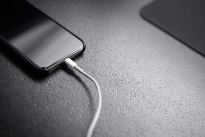 Close-up of a smartphone on a textured surface with a white charging cable connected to its bottom edge.