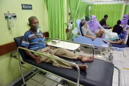 Older man on a hospital bed with an oxygen mask and IV line, in a ward with green curtains and other patients nearby.