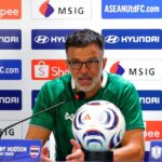 Man in a green jersey speaks at a press conference with a soccer ball on the table and sponsor banners behind him.