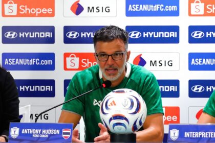 Man in a green jersey speaks at a press conference with a soccer ball on the table and sponsor banners behind him.