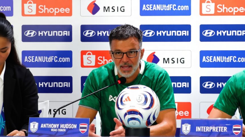 Man in a green jersey speaks at a press conference with a soccer ball on the table and sponsor banners behind him.