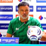 Head coach Anthony Hudson sits at a press conference table in a green shirt, with a football and microphone, sponsor backdrop behind him.