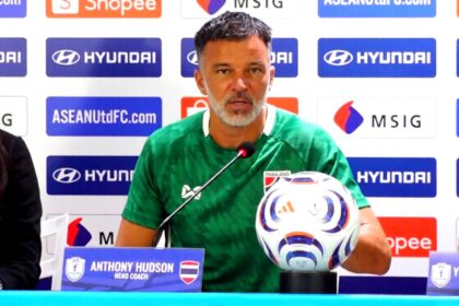 Head coach Anthony Hudson sits at a press conference table in a green shirt, with a football and microphone, sponsor backdrop behind him.