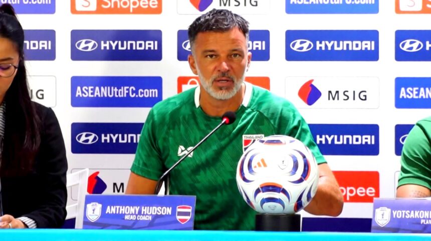 Head coach Anthony Hudson sits at a press conference table in a green shirt, with a football and microphone, sponsor backdrop behind him.