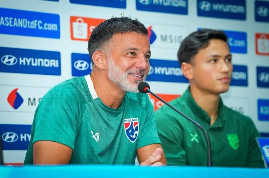 Two men in green jerseys sit at a press conference table, one speaking into a microphone with sponsor banners behind them.