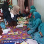 Group of women in turquoise outfits sitting on a colorful floor mat, exchanging papers.