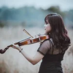 Woman playing a violin in an open field, wearing a black dress, with a soft, blurred background.