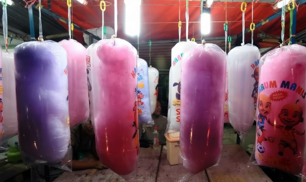 Rows of large purple and pink cotton candy cones wrapped in plastic, hanging at a market stall.