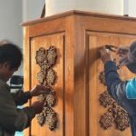 Two people attaching decorative star-shaped wooden inlays to a tall wooden cabinet or lectern, using tools on the panels.