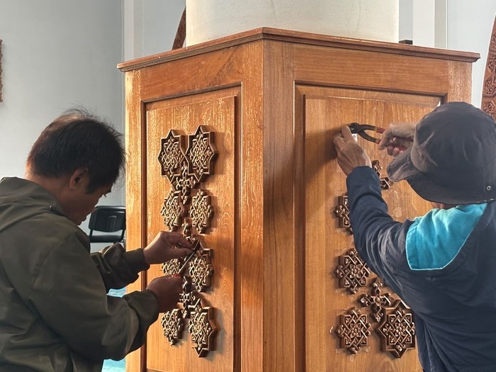 Two people attaching decorative star-shaped wooden inlays to a tall wooden cabinet or lectern, using tools on the panels.