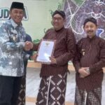 Four men in traditional Indonesian batik and peci on a stage during an award ceremony; two shake hands while one holds a document.