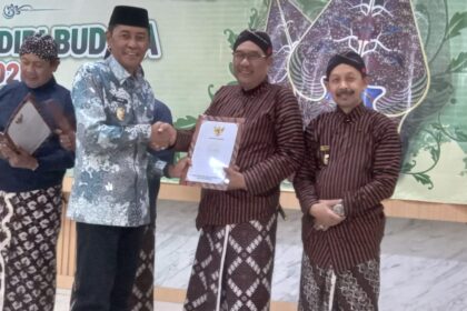 Four men in traditional Indonesian batik and peci on a stage during an award ceremony; two shake hands while one holds a document.
