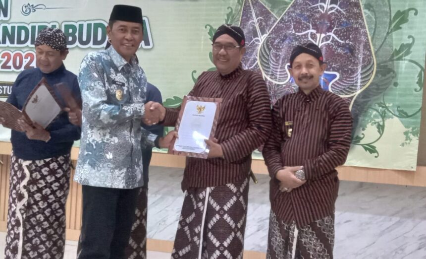 Four men in traditional Indonesian batik and peci on a stage during an award ceremony; two shake hands while one holds a document.