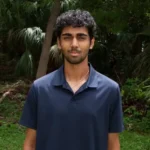 Young man with dark hair and light beard wearing a navy polo shirt stands outdoors in a tropical garden with palm trees behind him.