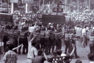 Riot police in helmets and shields block a city street as protesters gather; an armored vehicle sits behind them.
