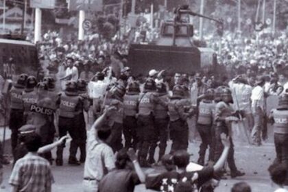 Riot police in helmets and shields block a city street as protesters gather; an armored vehicle sits behind them.