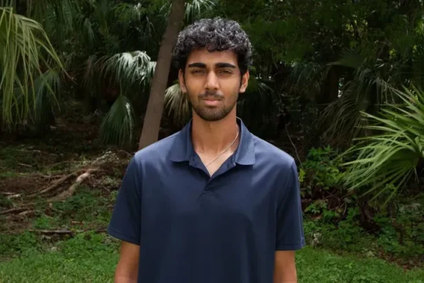 Young man with dark hair and light beard wearing a navy polo shirt stands outdoors in a tropical garden with palm trees behind him.