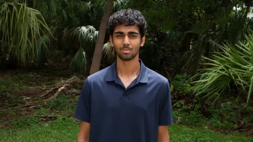 Young man with dark hair and light beard wearing a navy polo shirt stands outdoors in a tropical garden with palm trees behind him.