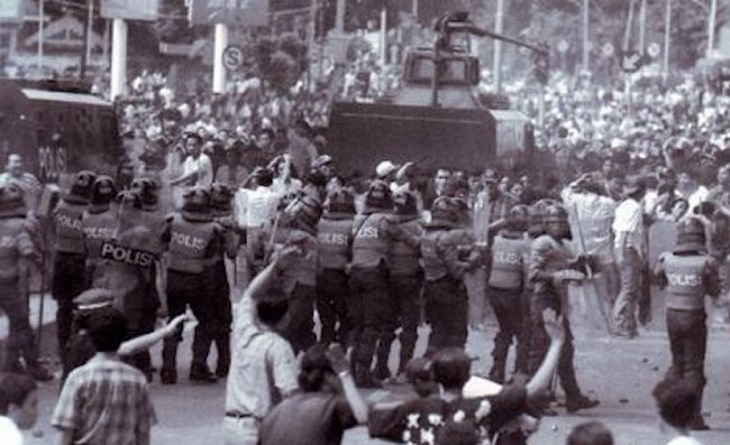 Riot police in helmets and shields block a city street as protesters gather; an armored vehicle sits behind them.