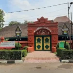 Red ornate gateway entrance to a compound with green-and-yellow double doors and banners along the fence.