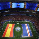 Aerial view of a packed stadium with oversized LED screens showing a trophy and bold graphics, and long national flag banners along the field for a FIFA World Cup event.