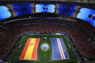 Aerial view of a packed stadium with oversized LED screens showing a trophy and bold graphics, and long national flag banners along the field for a FIFA World Cup event.