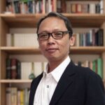 Professional man wearing glasses in a black blazer and white shirt standing in front of book-filled shelves.