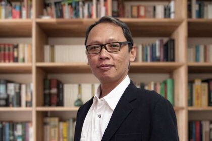 Professional man wearing glasses in a black blazer and white shirt standing in front of book-filled shelves.