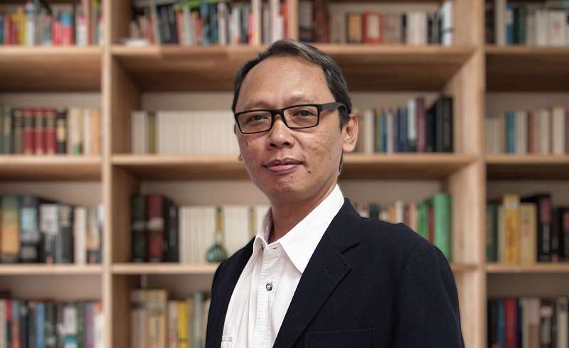 auA - Mabur.co Professional man wearing glasses in a black blazer and white shirt standing in front of book-filled shelves.