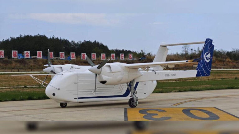 White twin-engine turboprop airplane parked on a taxiway with blue-tail livery and 'HH-100' on the fuselage, banners in the background.