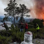 Four men drag a long hose toward a large brush fire in a hilly, smoky landscape as flames rise in the background.] , [Volunteers work together to fight a forest fire, hauling a water hose past a milestone marker.] , [Fire rages in the hills while a group of men pulls a long hose toward the flames, one wearing a high-visibility vest.] , [Smoky hillside fire with people guiding a hose toward the blaze near a roadside marker.]