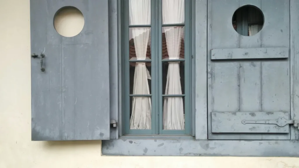 axA - Mabur.co Blue-gray wooden window with two panes and white curtains, flanked by circular cutouts in the shutters against a pale-yellow wall.