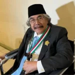 Elderly man in a wheelchair wearing a suit, tie, black hat, and a green‑white‑red ceremonial sash with a gold badge, smiling at the camera.