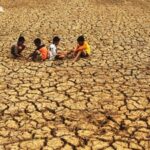Five children sit on parched, cracked earth in a drought-stricken landscape with a dusty horizon periphery.