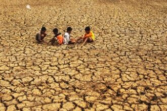 Five children sit on parched, cracked earth in a drought-stricken landscape with a dusty horizon periphery.