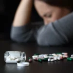 Person sits with head down at a table; an open bottle spills pills onto the surface, signaling distress.