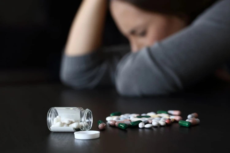 Person sits with head down at a table; an open bottle spills pills onto the surface, signaling distress.