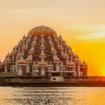 Sunset over a large lotus-shaped temple complex with orange domes by a calm river.