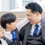 Adult man in a gray suit and blue tie hugs a young boy in a matching vest on outdoor stairs, smiling at each other.