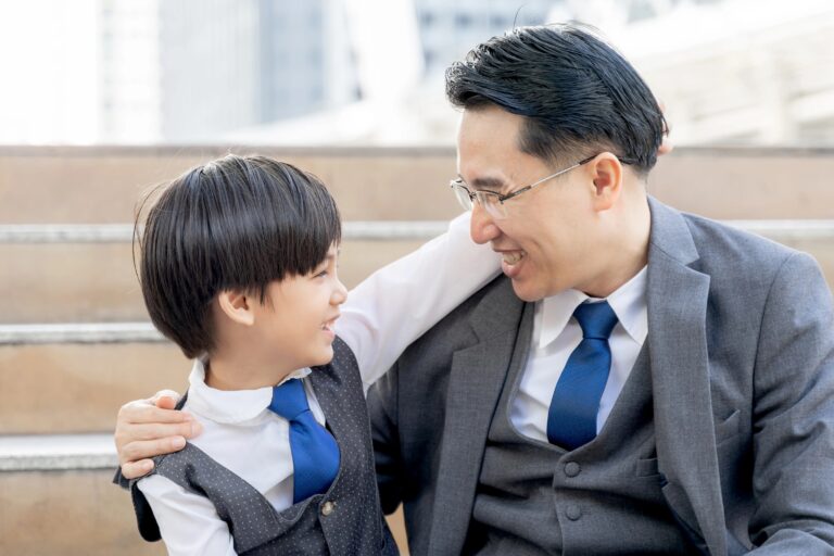 Adult man in a gray suit and blue tie hugs a young boy in a matching vest on outdoor stairs, smiling at each other.