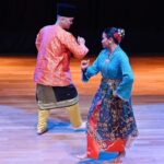 Two dancers in colorful traditional costumes perform a folk dance on a wooden stage.