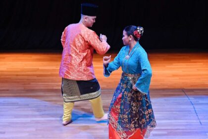 Two dancers in colorful traditional costumes perform a folk dance on a wooden stage.