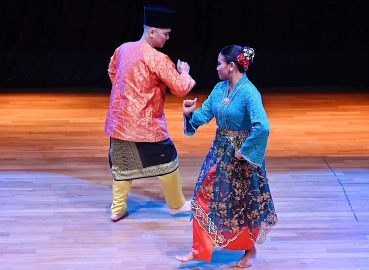 Two dancers in colorful traditional costumes perform a folk dance on a wooden stage.