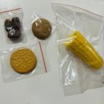 Assorted snacks in plastic bags: dates, a round cookie, a sandwich biscuit, and a corn cob.