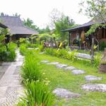 Tropical garden path with stepping stones, green lawn, and wooden cottages on either side.