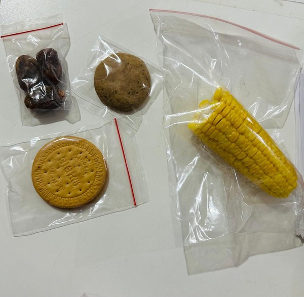 Assorted snacks in plastic bags: dates, a round cookie, a sandwich biscuit, and a corn cob.