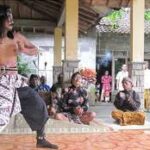 Two male performers in traditional loose pants dance on a raised platform as a small audience watches outdoors under a pavilion.