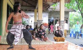 Two male performers in traditional loose pants dance on a raised platform as a small audience watches outdoors under a pavilion.