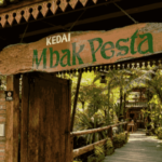 Wooden sign reading 'Kedai Mbak Pesta' over a rustic doorway leading to a leafy tropical cafe path.
