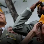 Three military personnel in dress uniforms salute while looking upward, two wearing yellow folded hats and one with sunglasses, during a ceremony.